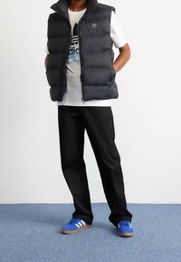 Svart pufferväst med hög krage, vit T-shirt med blå grafisk tryck, svarta byxor och blå Adidas-sneakers med vita ränder.