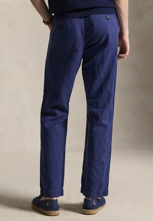 Mann trägt marineblaue gerade geschnittene Hose und passende Espadrilles, steht vor einem einfarbigen beigen Hintergrund.