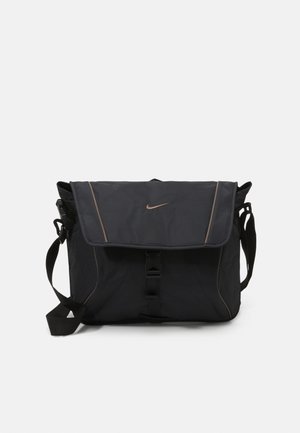 Sac messager en nylon noir avec rabat, fermeture à boucle et accents bronze ; comprend des poches latérales en mesh et une bandoulière rembourrée.
