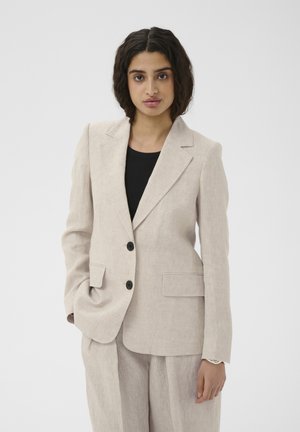 Ženska, ki nosi bež laneni blazer in ujemajoče hlače čez črno majico, stoji z eno roko v žepu pred enobarvnim ozadjem.