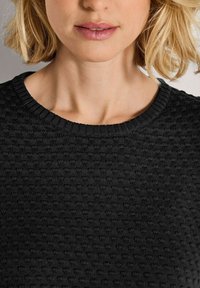 Eine blonde Frau trägt einen strukturierten schwarzen Strickpullover, gezeigt von Kinn bis zur oberen Brust vor einem neutralen Hintergrund.