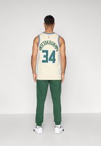 Camisa de basquetebol creme com detalhes azuis, apresentando "ANTETOKOUNMPO" e o número "34" nas costas. Usada com calças de fato verdes e sapatilhas brancas.
