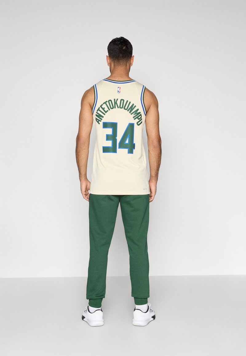 Maillot de basketball crème avec des bordures bleues, portant "ANTETOKOUNMPO" et le numéro "34" dans le dos. Porté avec un pantalon de jogging vert et des baskets blanches.