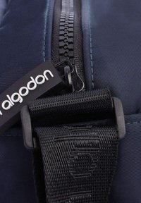 Primer plano de una bolsa de tela azul marino, con un zipper negro, correa tejida y una etiqueta de marca. La textura es suave con detalles de costura visibles.