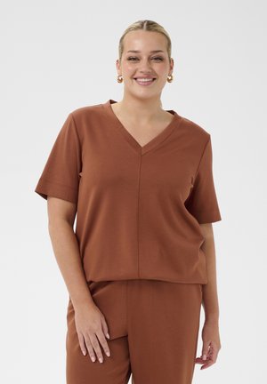 Femme souriante aux cheveux blonds attachés en queue de cheval basse, portant des boucles d'oreilles anneaux dorés, un haut marron à manches courtes et col en V ainsi qu'un pantalon assorti.