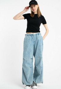 Młoda kobieta w czarnym krótkim topie, czapce w czarnym kolorze, szerokich jasnoniebieskich dżinsach oraz czarno-białych sneakersach, pozująca z jedną ręką na czapce, a drugą w kieszeni.