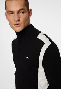 Svart pullover med hög krage, med vita detaljer på axlarna och halvdragkedja. Litet logotyp på bröstet. Slät textur.