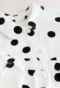 Tissu blanc à pois noirs, avec un col, des boutons-pression et une taille élastique sur un vêtement.