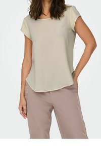 Blouse beige clair à manches courtes avec une coupe carrée et un col rond. Le tissu présente une texture lisse. Comprend des fentes latérales pour faciliter les mouvements.