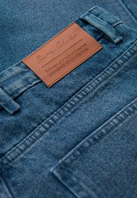 Jeans de denim con un lavado azul oscuro, que cuentan con un parche de cuero marrón en la cinturilla con texto en relieve y detalles de costura en naranja.