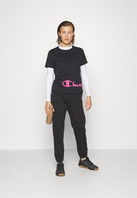 Camiseta negra de manga corta con logo rosa, superpuesta sobre mangas largas blancas, combinada con pantalones de chándal negros y zapatillas negras, sosteniendo una botella de agua marrón.