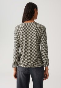 Vrouw die wegkijkt, gekleed in een langmouwtop met een zwart, wit en beige geometrisch kubuspatroon en donkergrijze denim jeans.