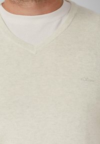 Lichtbeige V-halstrui van gebreid materiaal, met een subtiele textuur en een klein logo aan de voorkant links.