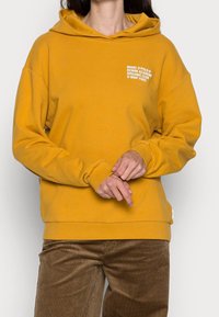 Sudadera con capucha color amarillo mostaza de corte holgado, con texto blanco en el pecho y puños acanalados. Combinada con pantalones de pana marrón.