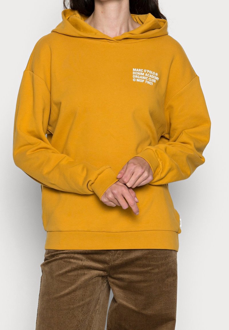 Sudadera con capucha color amarillo mostaza de corte holgado, con texto blanco en el pecho y puños acanalados. Combinada con pantalones de pana marrón.