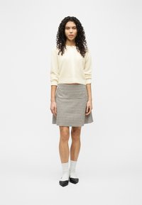 Γυναίκα με σγουρά μαλλιά, ντυμένη με κρεμ ζακέτα, καρό φούστα, λευκές κάλτσες και μαύρα παπούτσια με μυτερή μύτη, στέκεται μπροστά σε έναν απλό φόντο.