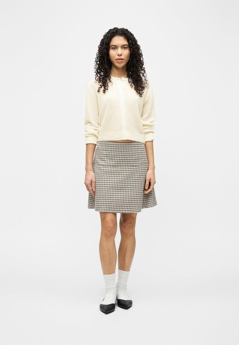 Γυναίκα με σγουρά μαλλιά, ντυμένη με κρεμ ζακέτα, καρό φούστα, λευκές κάλτσες και μαύρα παπούτσια με μυτερή μύτη, στέκεται μπροστά σε έναν απλό φόντο.