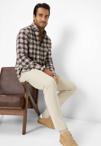 Geruite shirt in bruin, crème en marineblauw; doorgeknoopt, met een kraag. Lichtbeige broek en tan sneakers met witte accenten. Houten stoel.