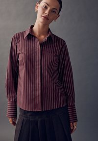 Bordó csíkos ing gombokkal, hosszú ujjakkal és hegyes gallérral, fehér függőleges csíkokkal és kontrasztos mandzsettákkal.