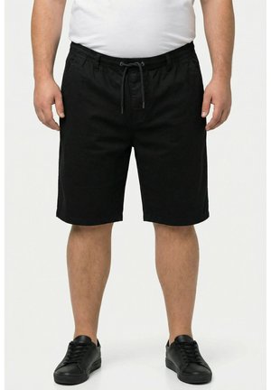 Uomo con pantaloncini neri con coulisse, sneakers nere e t-shirt bianca, in piedi davanti a uno sfondo semplice.