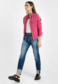 Felle roze denimjack met twee borstzakken, gedragen over een wit shirt. Blauwe jeans met een licht vervaagde afwerking en opgerolde manchetten, gecombineerd met zwarte sandalen.