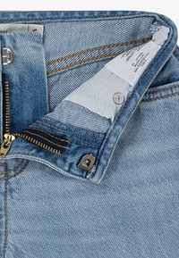 Světle modré džíny z denimu s upínáním na zip se zlatými detaily, oranžovým prošíváním a látkovou etiketou uvnitř pásu.