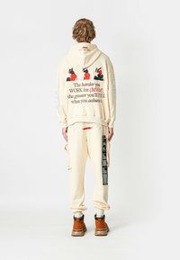 Persona vestida con sudadera crema y pantalones con gráfico y texto en rojo y negro, botas marrones, de pie, de espaldas, sobre fondo blanco.