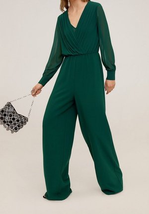 Une combinaison verte avec un décolleté en V profond, un corsage plissé, des manches longues en voile et des jambes larges. Associée à un sac à main noir à motif circulaire.