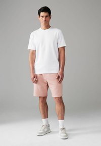 Witte katoenen t-shirt gecombineerd met roze shorts, met lichte sneakers en witte sokken, staand tegen een grijze achtergrond.