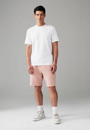 T-shirt en coton blanc associé à un short rose, complété par des baskets de couleur claire et des chaussettes blanches, se tenant contre un fond gris.