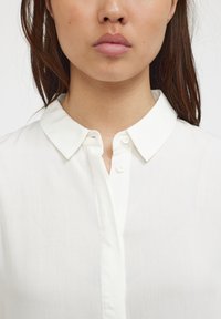 Camicia bianca con colletto classico, patta bottoni e texture liscia. Presenta un design pulito e semplice, senza motivi né decorazioni.