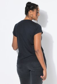 Mujer con cabello oscuro recogido en un moño, usando pendientes de aro, una camiseta negra y jeans negros, de pie y de frente a una pared blanca en un estudio.