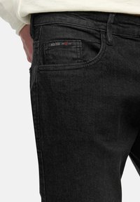 Jeans de denim negro con corte ajustado, que cuentan con un bolsillo trasero con una etiqueta de la marca y sutiles detalles de textura. Cierre de botón seguro.