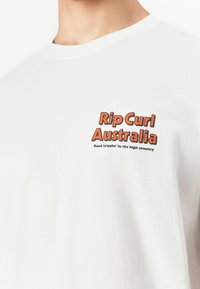 Hvit T-skjorte med oransje og svart "Rip Curl Australia" tekst og en mindre slagord "Road trippin' in the high country" på øvre bryst.