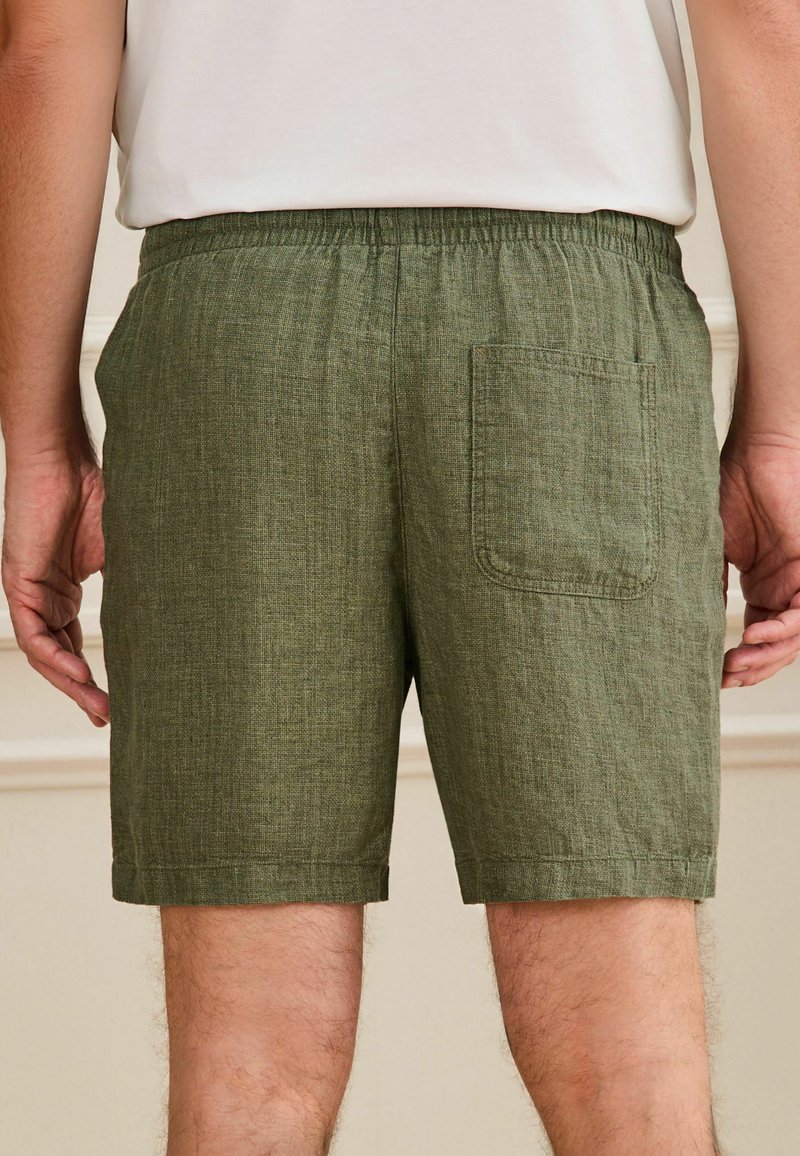 Homme portant un short en lin vert olive avec taille élastique et une poche arrière, debout, les bras détendus le long du corps.