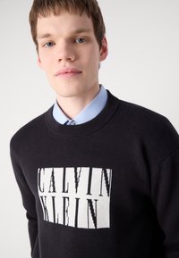 Black knitted sweater featuring a prominent white "CALVIN KLEIN" logo. The collar is ribbed, and the model wears a light blue shirt underneath.