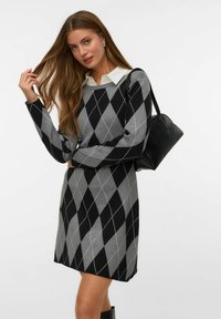 Donna che indossa un vestito a maglione argyle nero e grigio con una camicia colletto bianca, tiene in mano una borsa nera e tocca i suoi lunghi capelli castani.