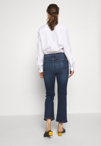 Vaqueros de tiro alto en azul con pierna acampanada, que cuentan con bolsillos traseros y detalles de costura, combinados con una camisa blanca de manga larga y tacones negros.
