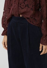 Blouse en dentelle bourgogne avec un motif floral, associée à un pantalon en velours côtelé bleu marine à rayures verticales et à coupe décontractée.