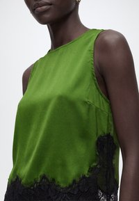 Femme portant un top sans manches en satin vert avec une bordure en dentelle noire à l'ourlet sur un fond clair uni.