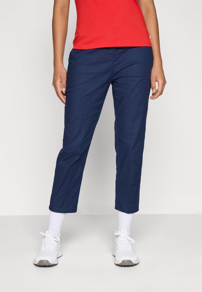 Persona in pantaloni cropped blu navy, calze bianche, sneaker bianche e una maglietta rossa, in piedi contro uno sfondo chiaro e uniforme.