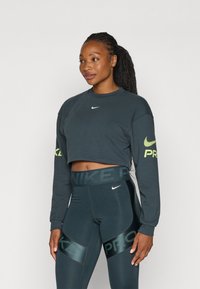 Mørkegrå cropped sweatshirt med lange ærmer og Nike-logo; højtaljede leggings med dæmpede grønne mønstre og brandaccentueringer.