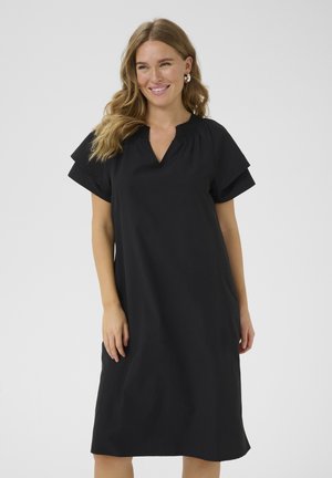 Donna sorridente con capelli biondi ondulati indossa un abito nero fluido fino al ginocchio con maniche corte a strati e colletto a V arricciato.