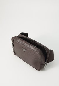 Bolso cruzado de cuero marrón texturizado con herrajes de plata, cierre de cremallera y acento de logo. Forma rectangular compacta con correa ajustable.