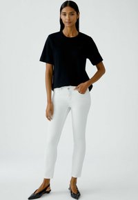 Zwart T-shirt met korte mouwen gecombineerd met witte strakke jeans. Het model staat rechtop, met een hand in de zak en draagt zwarte schoenen.