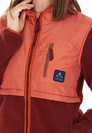 Fleece- en nylonjack in oranje en bordeauxrood met een ritsluiting, borstzak en logo-patch. Heeft een hoge kraag en een gestructureerde stof.