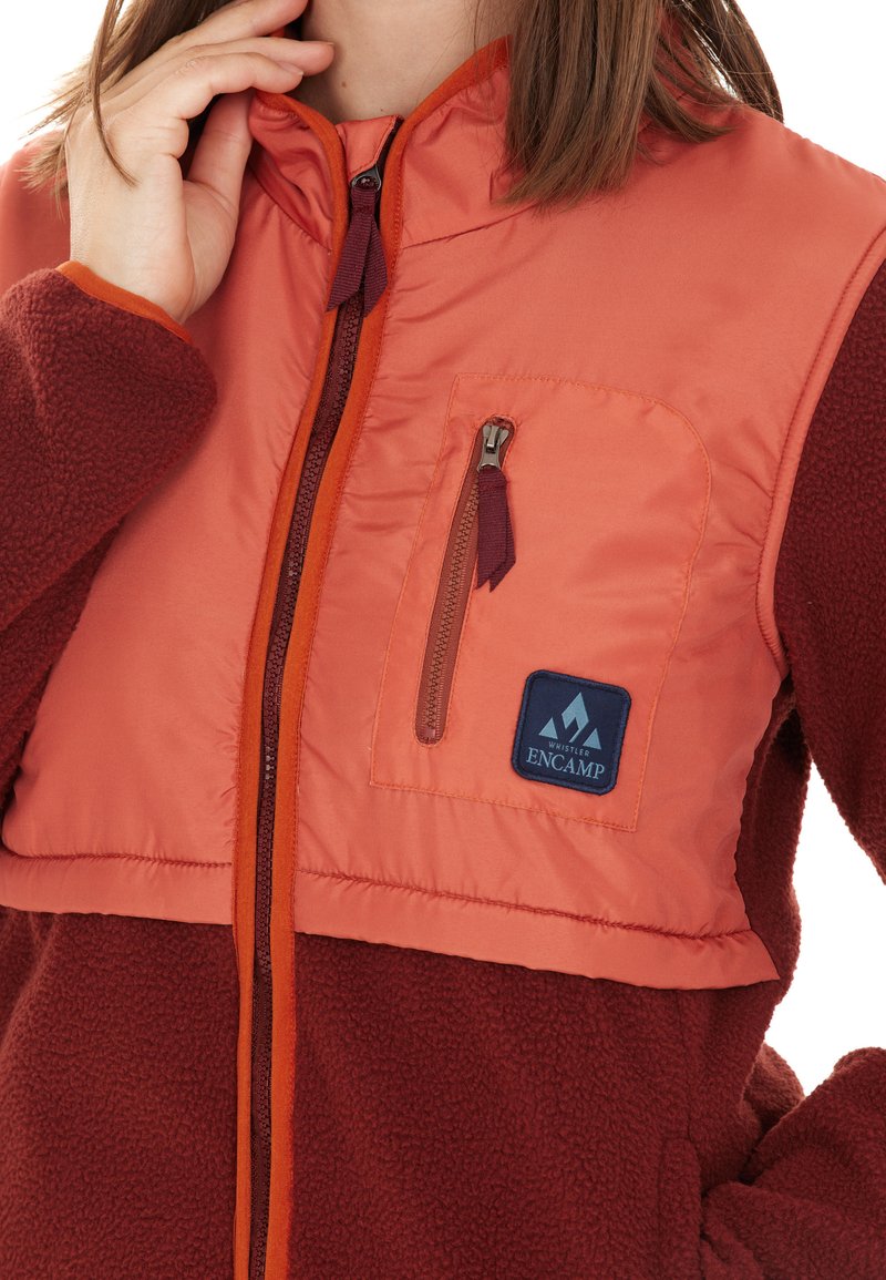 Fleece- en nylonjack in oranje en bordeauxrood met een ritsluiting, borstzak en logo-patch. Heeft een hoge kraag en een gestructureerde stof.