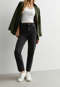 Zwarte high-waisted jeans met een versleten kniedetail, gecombineerd met een witte tanktop en een donkergroene oversized blazer, en witte sneakers.