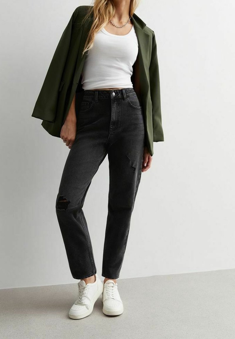 Zwarte high-waisted jeans met een versleten kniedetail, gecombineerd met een witte tanktop en een donkergroene oversized blazer, en witte sneakers.