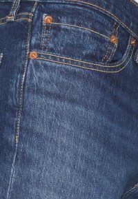 Close-up of blue denim jeans showing front pocket with copper rivets and yellow stitching details.